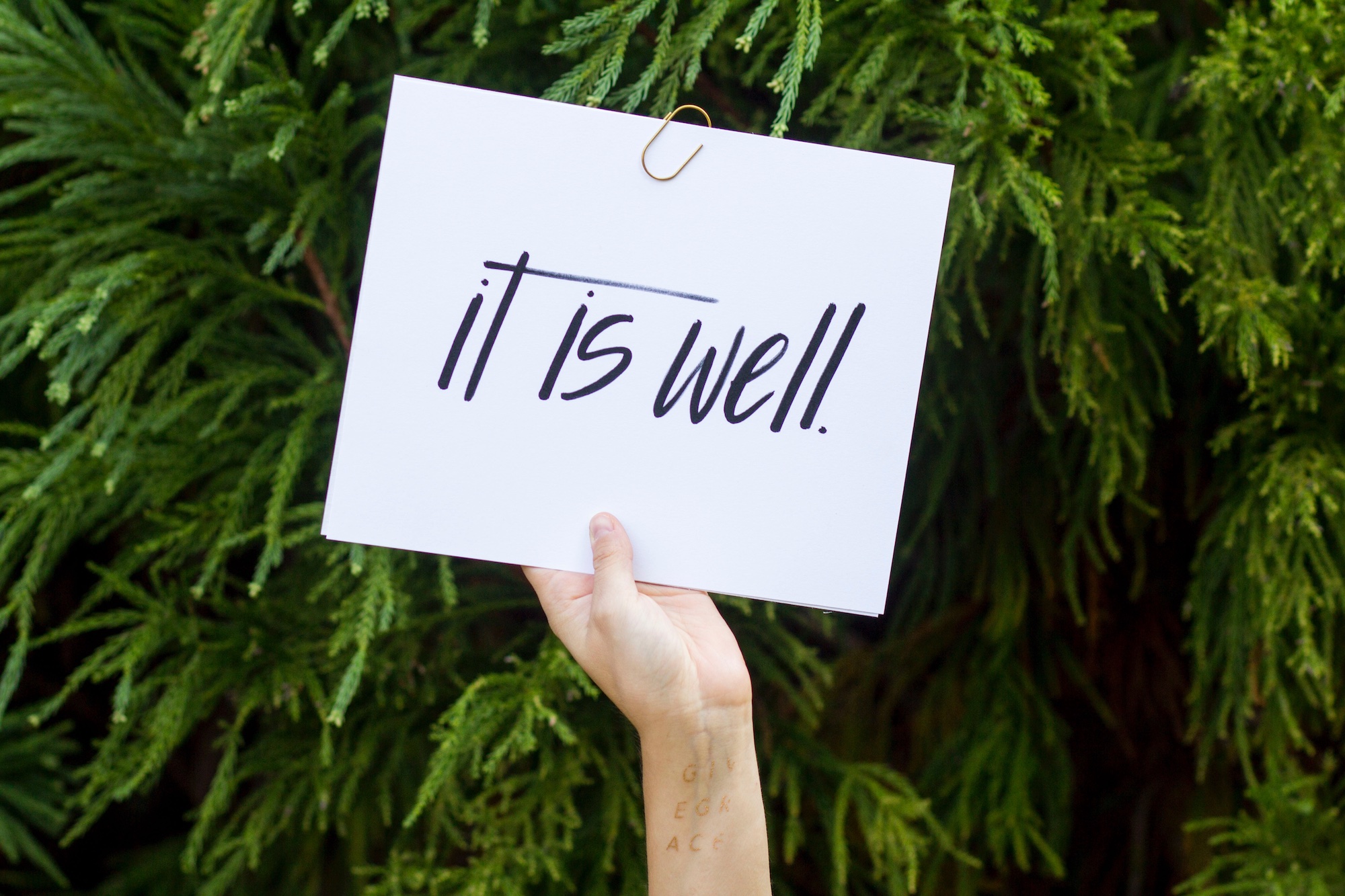 Hand holding a sign in front of a green tree that reads, "it is well."