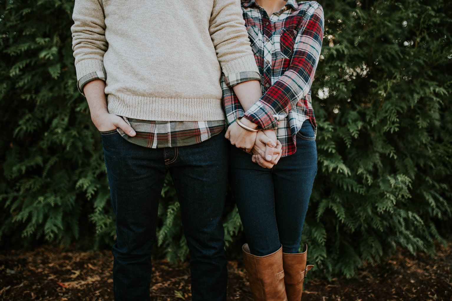 Shot of a couple from the chest down holding hands in front of a large green bush