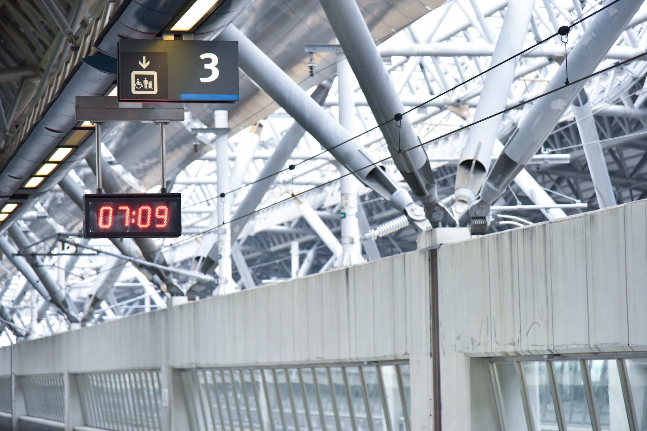 Digital clock displaying "07:09" in red print hanging from the ceiling in a white subway station.