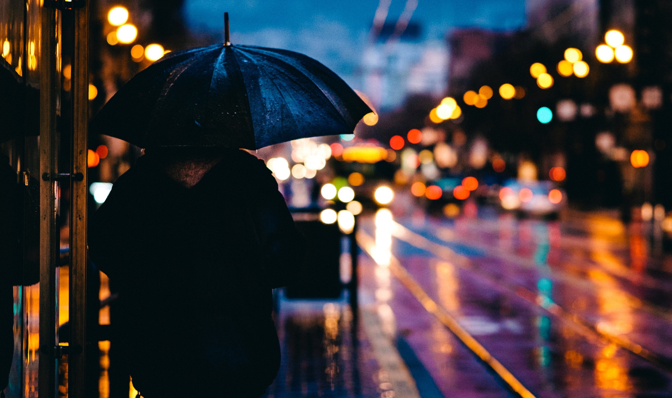 Cloaked man under an umbrella on a sidewalk near bright lights