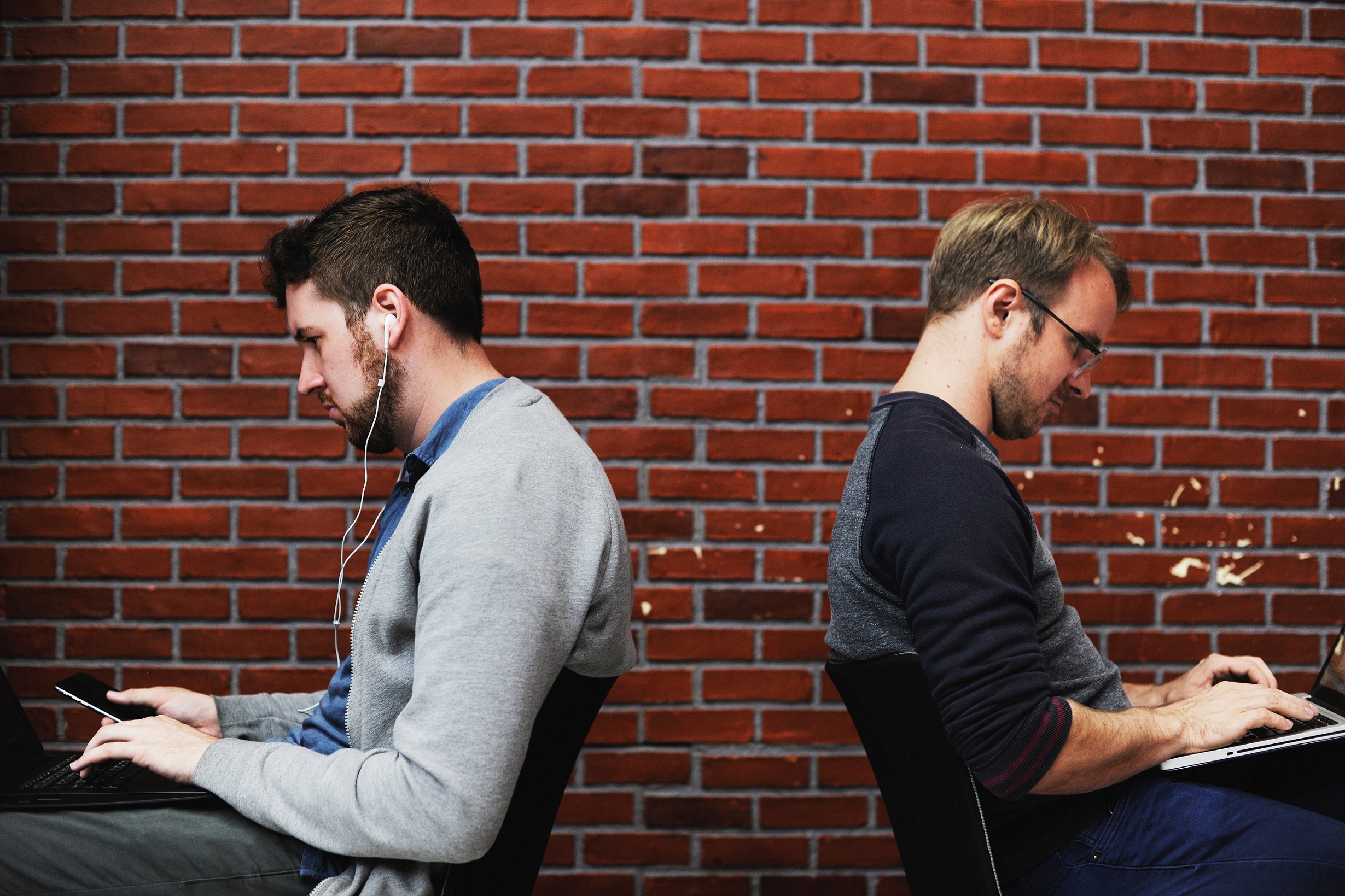 Two men sitting in chairs back to back while using a phone and laptop
