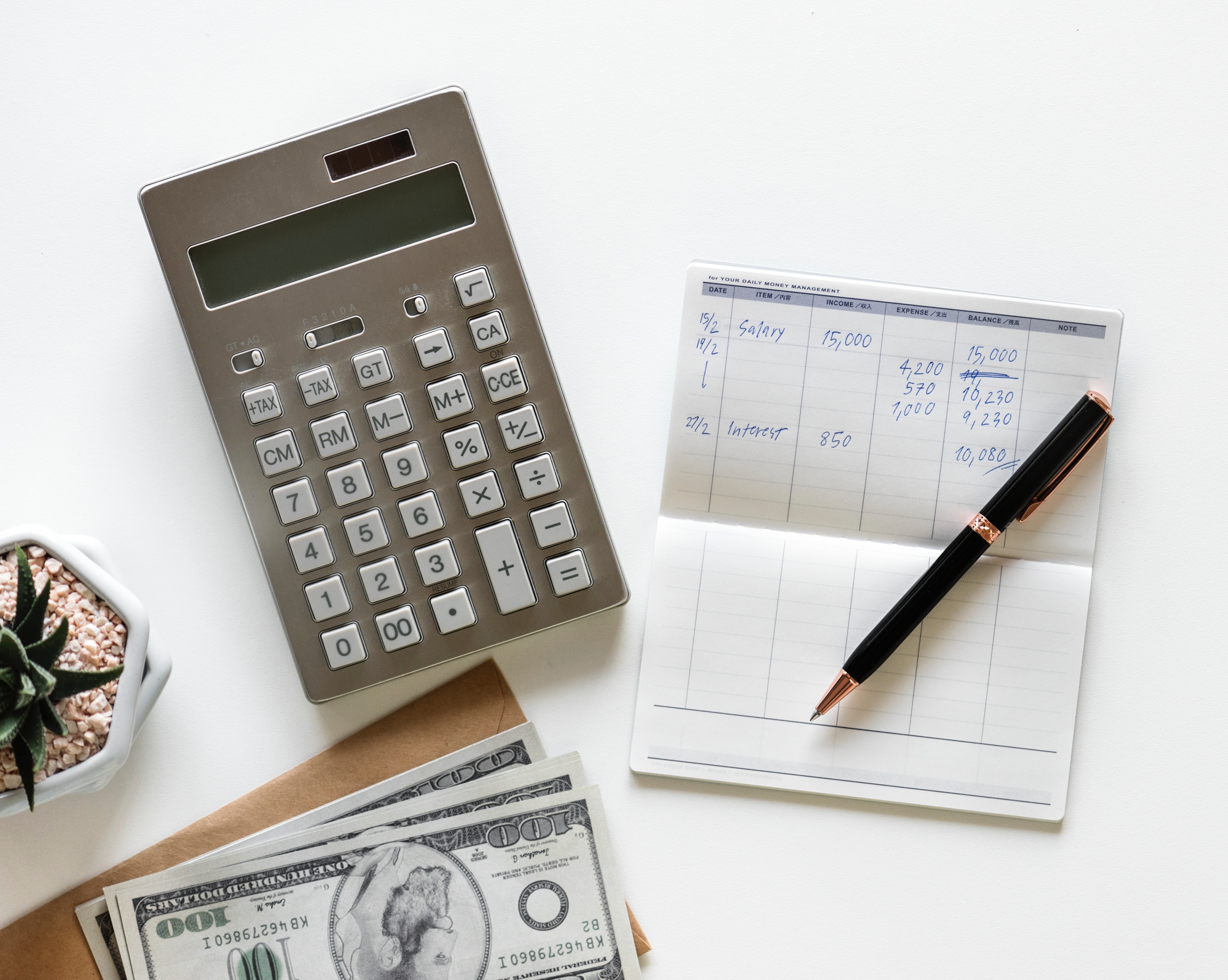 Calculator, pen on top of a checkbook, and a small stack of $100 bills.