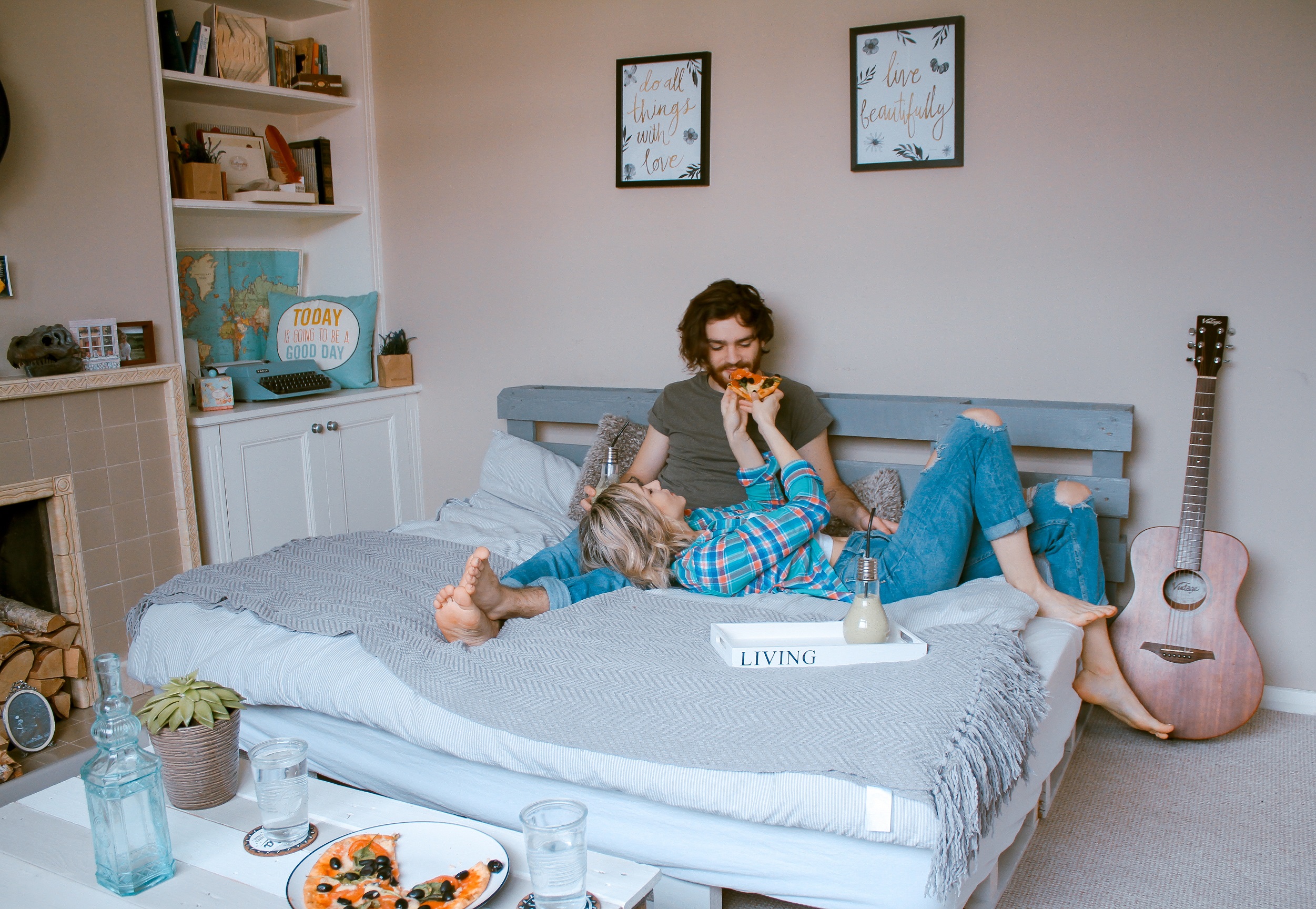 Man and woman relaxing on a pastel comforter; woman feeding man pizza while lying her head on his legs.