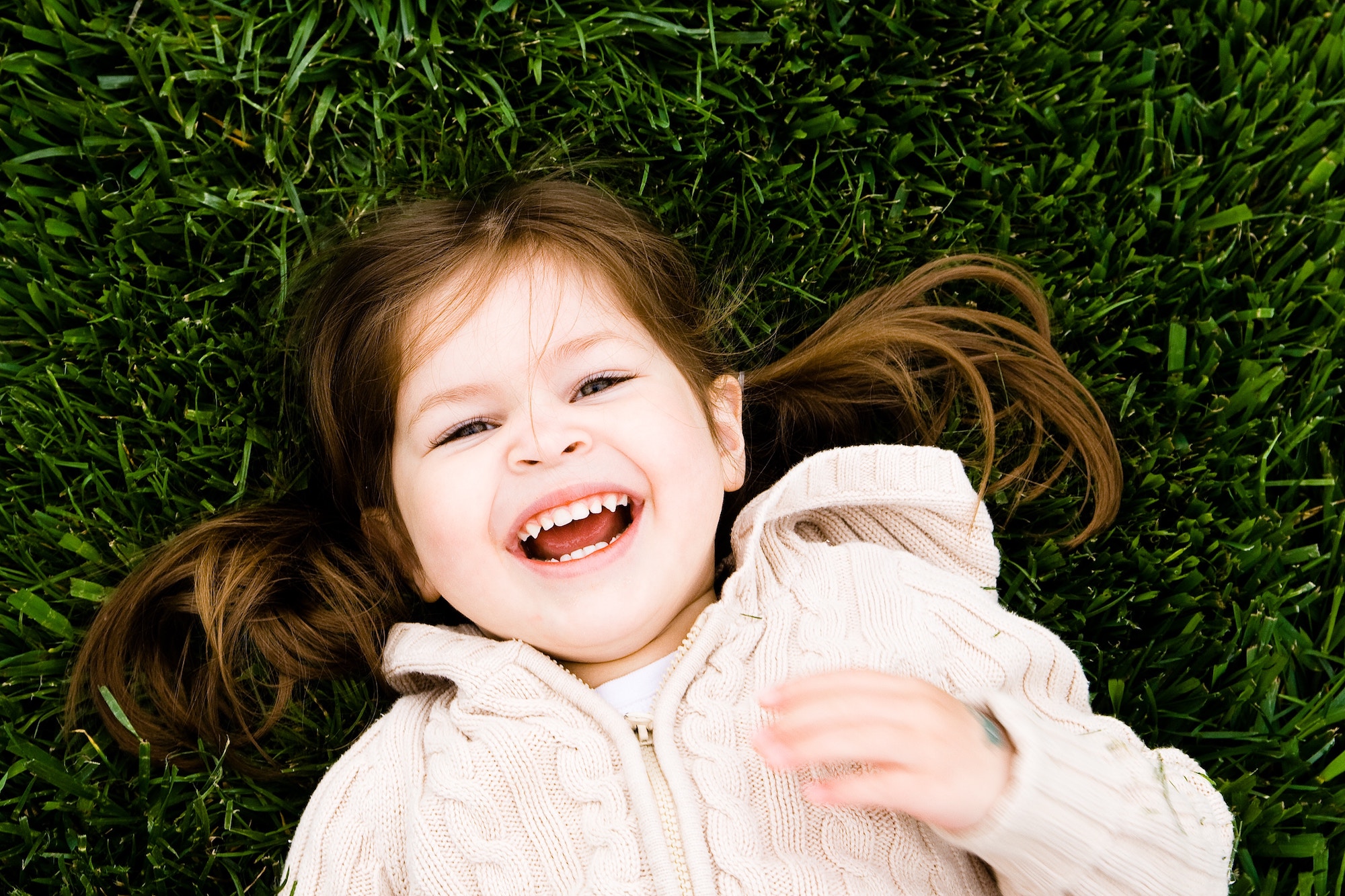 Little girl laying on grass in a pink jacket, smiling as the camera captures her from above.