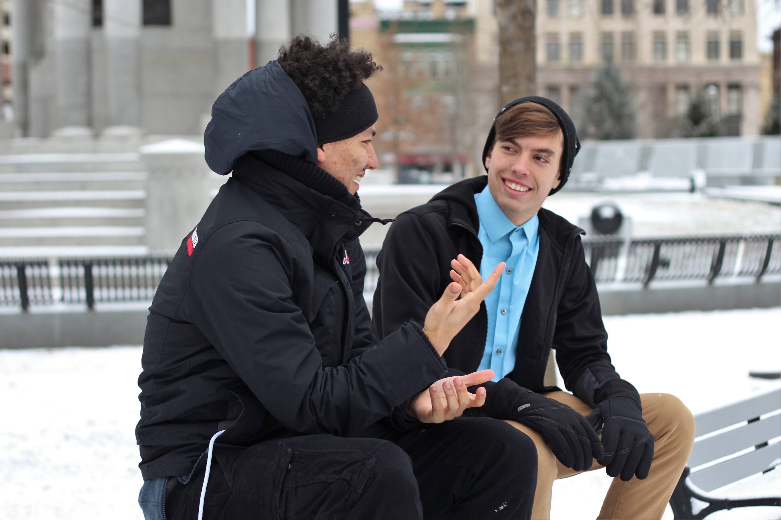 Two guys sitting on a park bench in the snow having a conversation.