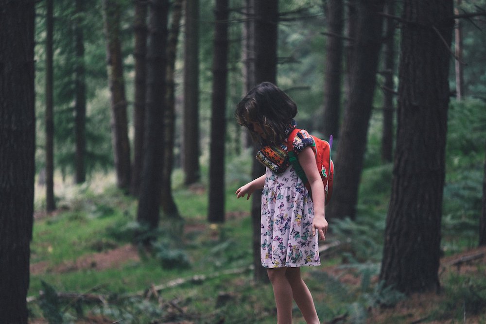 Young girl walking in the woods with a red backpack on, looking downward