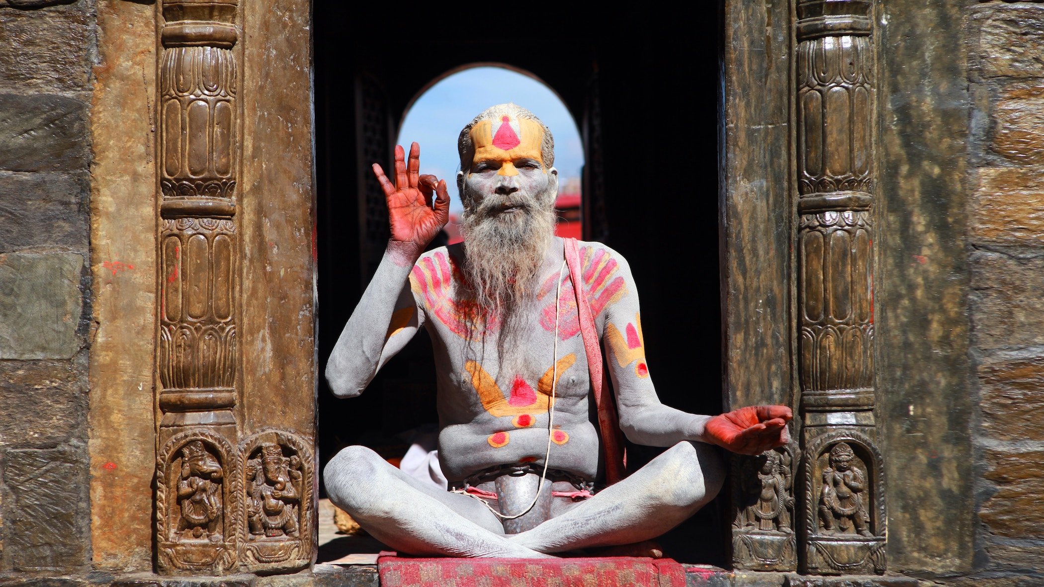 Man covered in body paint meditating in the sun, the fingers on his right hand forming an 