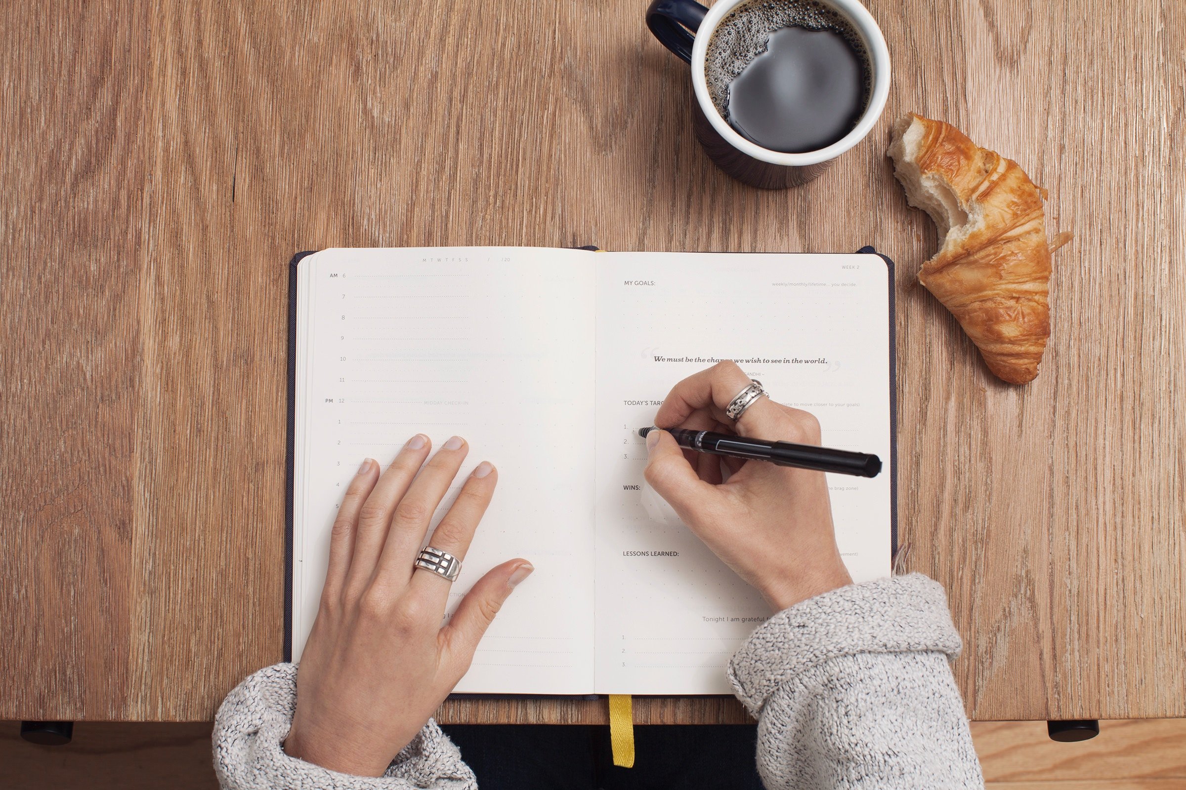 Image of coffee cup, croissant with a bite taken out of it, a planner, and a woman with a pen beginning to write in the planner.