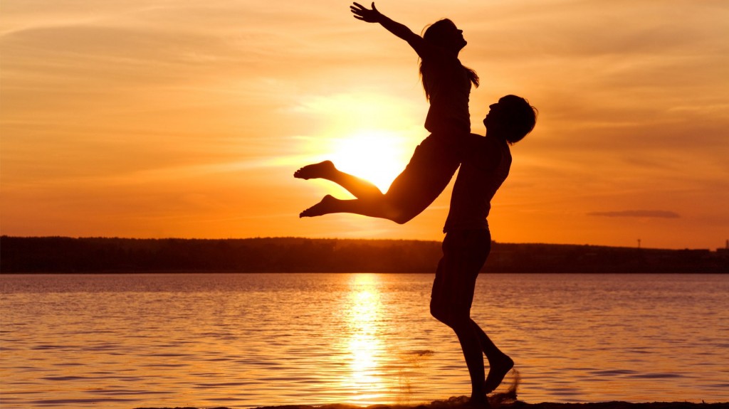 Silhouette of a woman jumping into man's arms in front of an ocean sunset