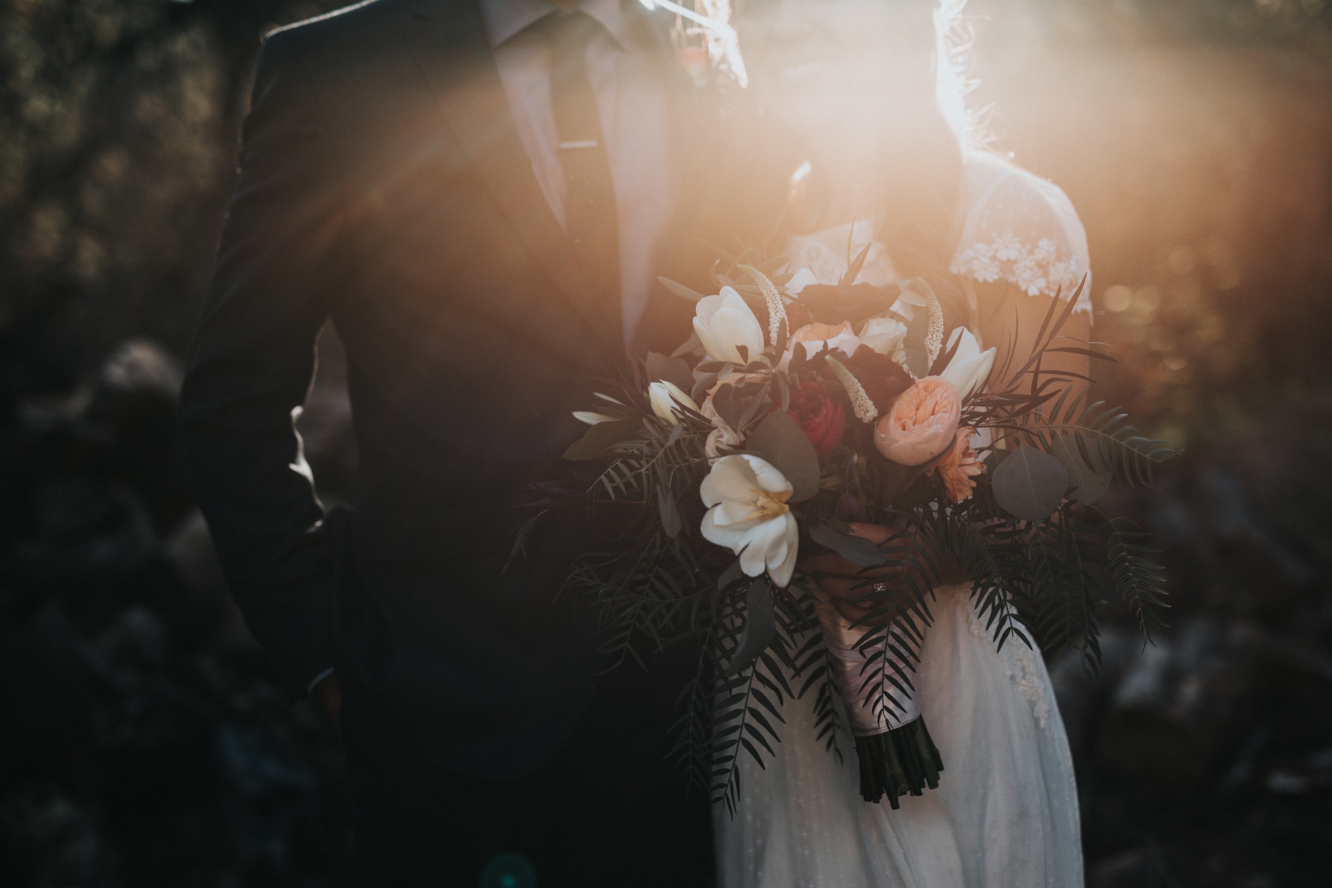 Slightly darkened image of a man in a tux and woman in a wedding dress holding a bouquet of flowers shot from the shoulders down.