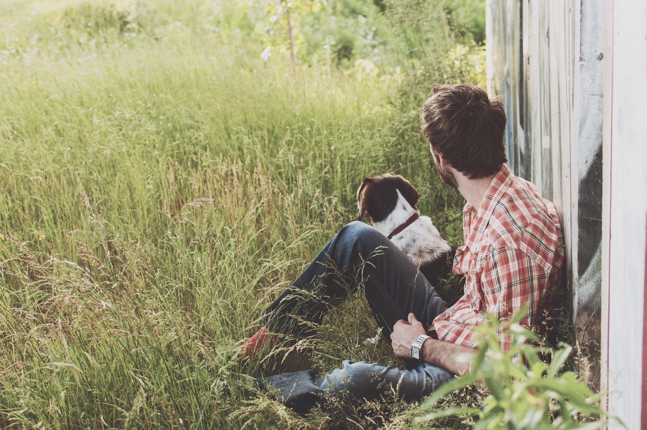 Man and dog sitting against the side of a barn in high grass