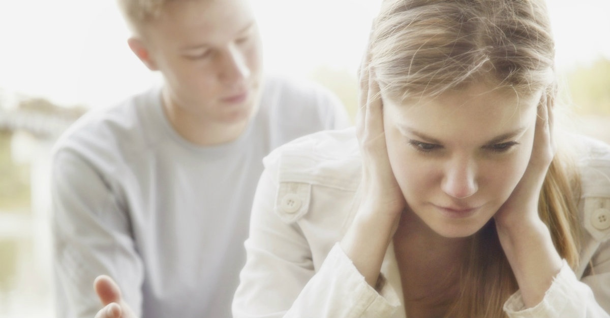 Man in background trying to talk to his spouse, but she has her hands over her ears and eyes closed.